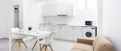 A modern kitchen with white cabinets, a sink, and a microwave. There is a dining table with chairs and a beige sofa.