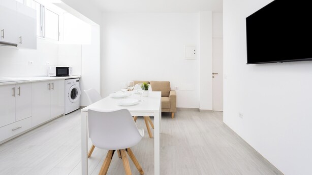 Una cocina moderna con armarios blancos, una mesa de comedor con sillas, lavadora y televisor de pantalla plana montado en la pared.