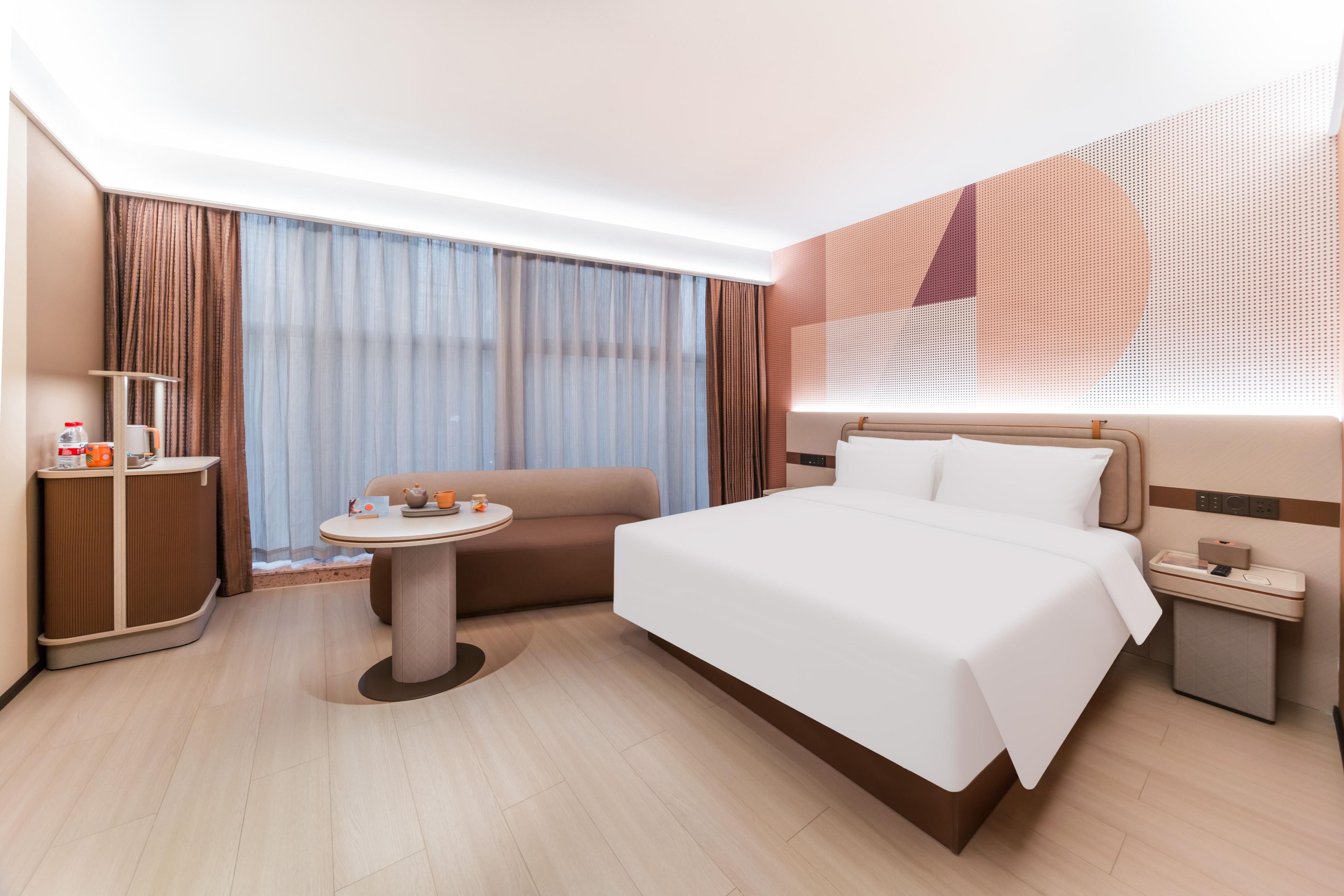 Et moderne hotelværelse med en stor seng, et lille rundt bord, en sofa, et skrivebord med lampe og et stort vindue med gardiner.