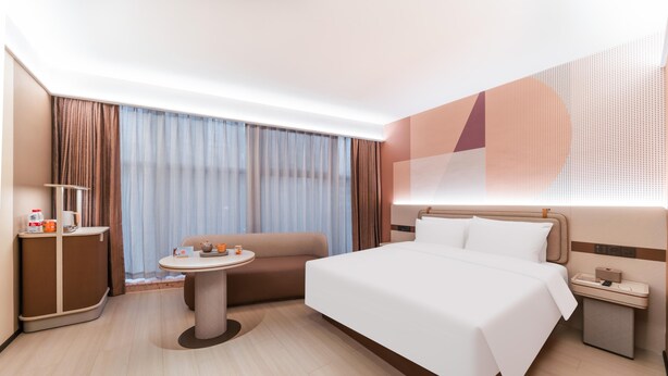 A modern hotel room with a large bed, a small round table, a sofa, a desk with a lamp, and a large window with curtains. - Orange Hotel Guangzhou Tianhe Sports Center (Guangzhou)