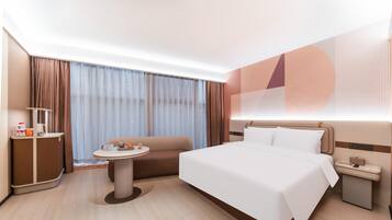 Una habitación de hotel moderna con una cama grande, una mesita redonda pequeña, un sofá, un escritorio con lámpara y un ventanal con cortinas.