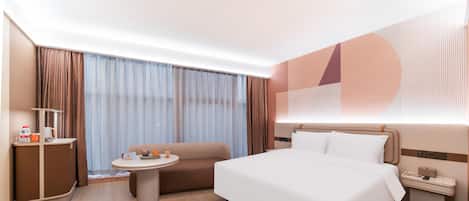 Una habitación de hotel moderna con una cama grande, una mesita redonda pequeña, un sofá, un escritorio con lámpara y un ventanal con cortinas.