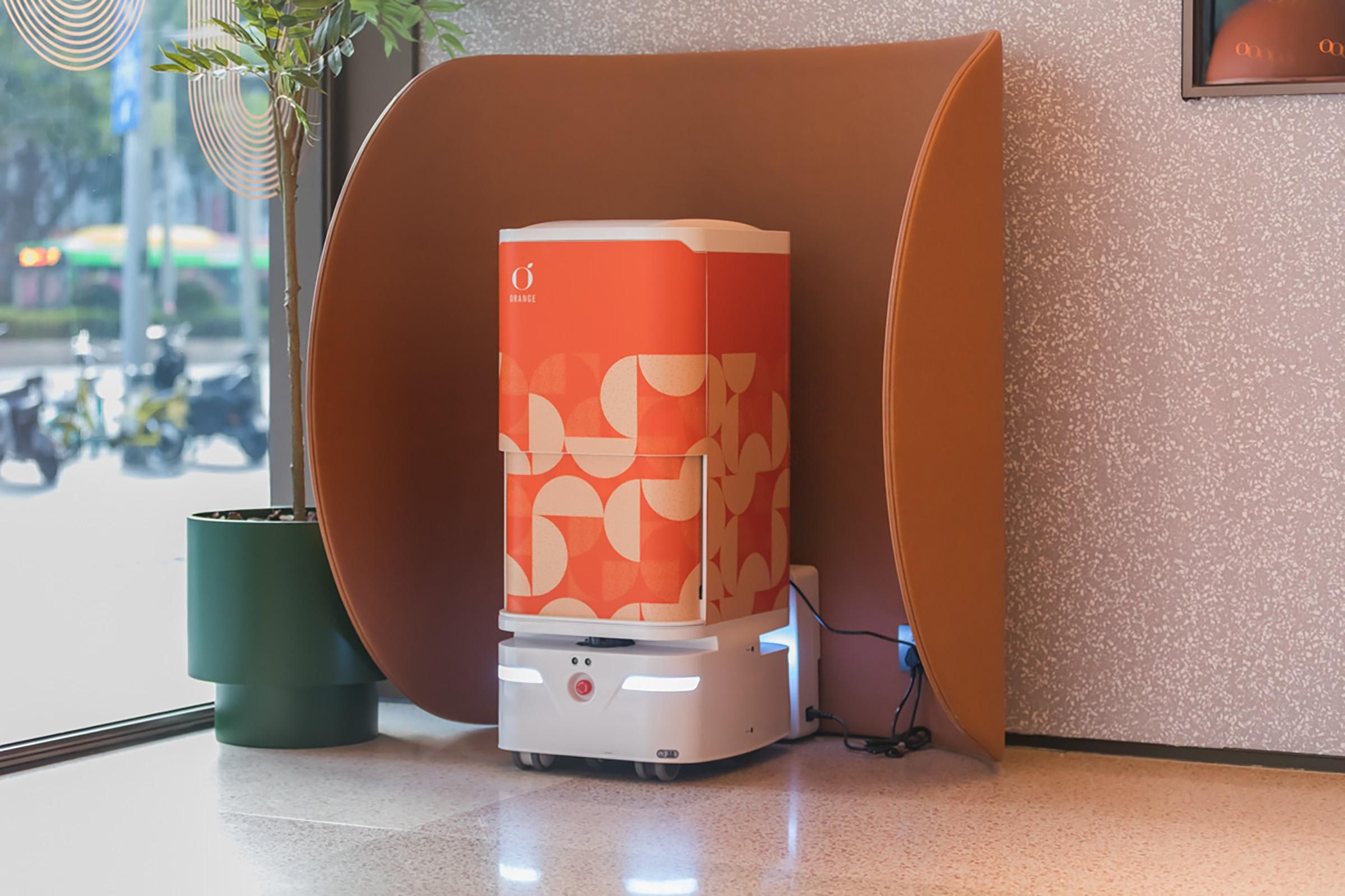 Una cafetera compacta y moderna con un diseño distintivo naranja y blanco, sobre un piso pulido cerca de una ventana.