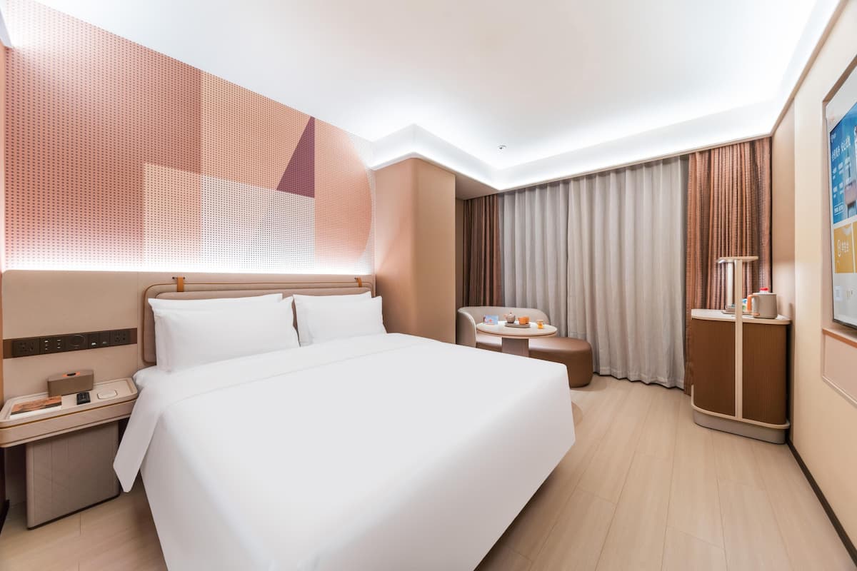 A hotel room with a large bed, a desk with a telephone, a chair, a small table with a cup, and a cabinet.