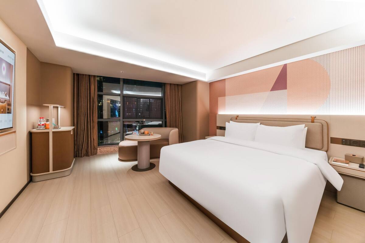 A modern hotel room with a large bed, a small round table, a sofa, and a desk with a lamp.