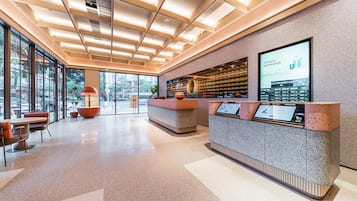 Un área de recepción moderna con un muro de vidrio, una mesa de recepción y una zona de estar con sillas naranjas.