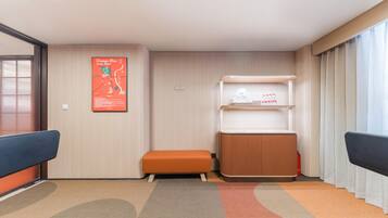 A hotel room with a large window, a poster on the wall, a bench, a small table, and a carpeted floor.