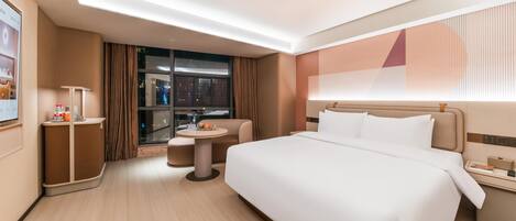 A modern hotel room with a large bed, a small round table, a sofa, and a desk with a lamp.