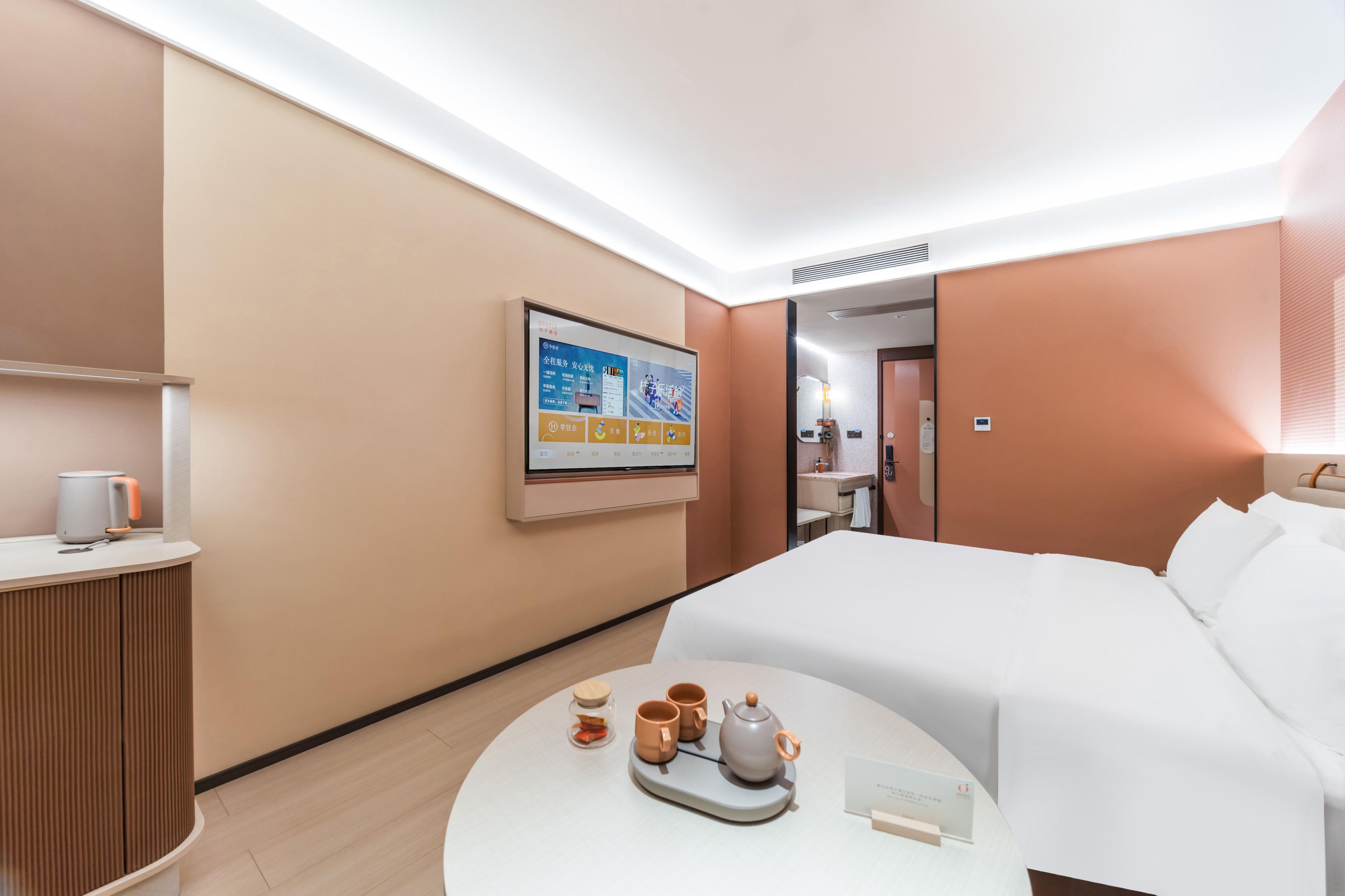 Quarto de hotel com uma cama, uma mesinha de cabeceira com chaleira e chávenas, um quadro na parede e uma secretária com uma lâmpada.