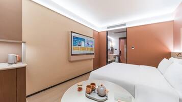 Quarto de hotel com uma cama, uma mesinha de cabeceira com chaleira e chávenas, um quadro na parede e uma secretária com uma lâmpada.