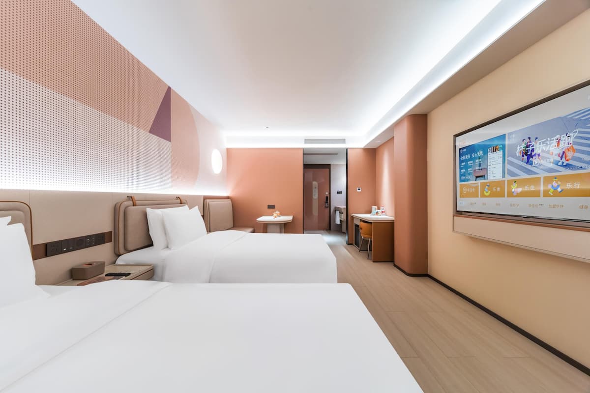 A modern hotel room with two beds, a small table, a desk, and a large TV screen displaying a menu.