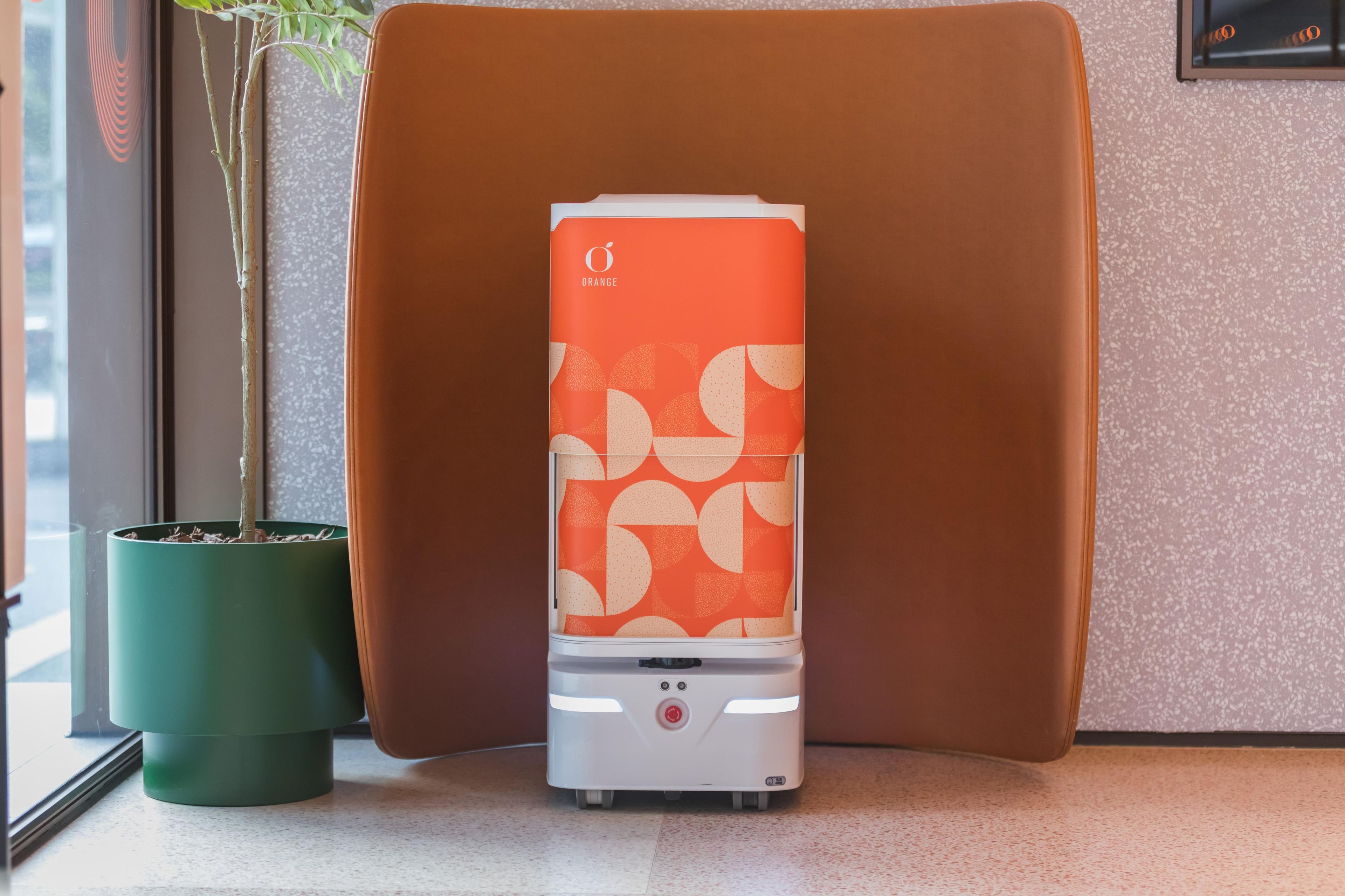 Un dispositivo compacto, de diseño naranja y blanco, posiblemente un calentador portátil o purificador de aire, situado sobre un suelo moteado junto a una planta en maceta verde y una silla marrón.