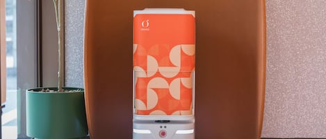 Un dispositivo compacto, de diseño naranja y blanco, posiblemente un calentador portátil o purificador de aire, situado sobre un suelo moteado junto a una planta en maceta verde y una silla marrón.