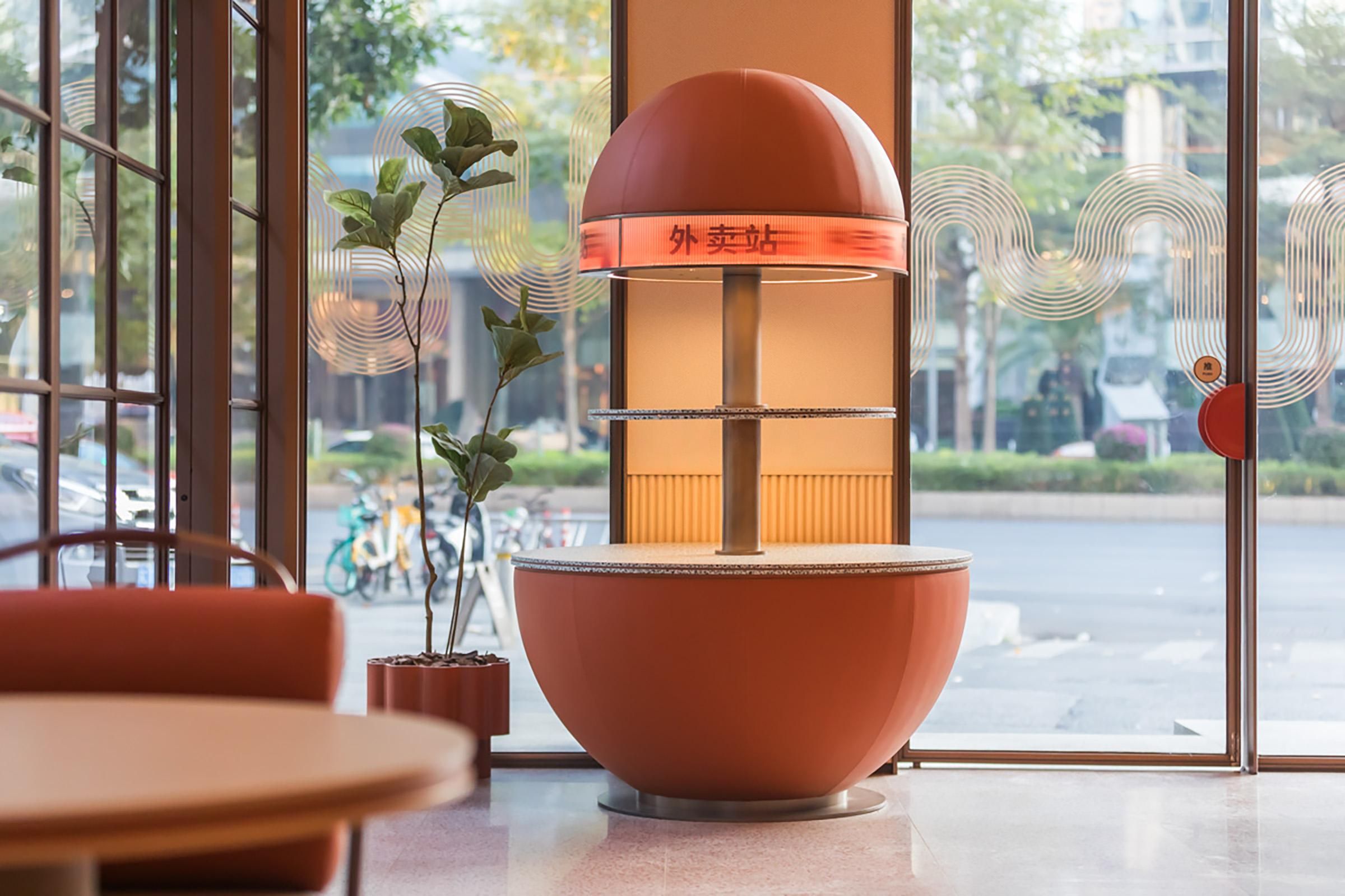 Un interior moderno con una lámpara naranja grande, plantas en macetas y vistas de la calle a través de grandes ventanales.