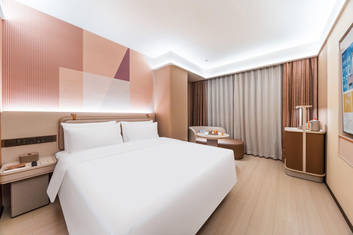 A hotel room with a large bed, a desk with a telephone, a chair, a small table with a cup, and a cabinet.