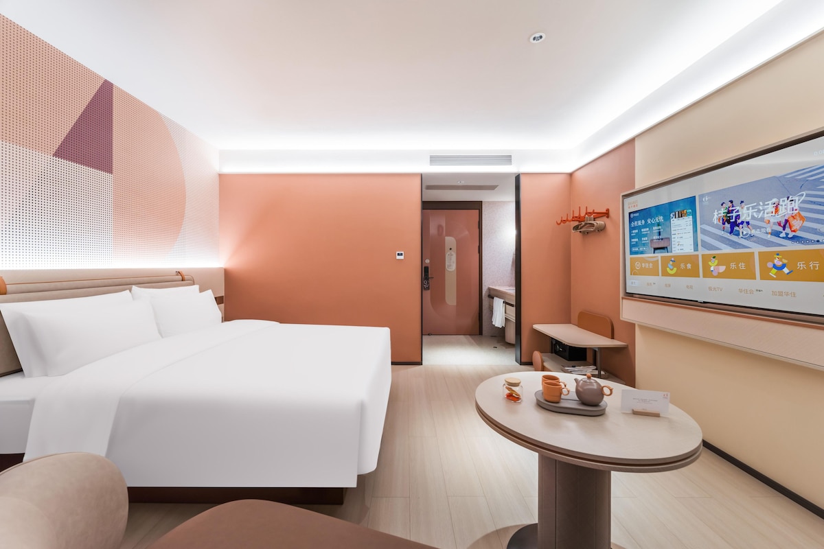 A hotel room with a bed, a round table with a teapot, a chair, a small table, and a wall-mounted TV displaying a menu.