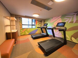 A hotel room with two treadmills, a bench, a desk, and a large window. - Orange Hotel Guangzhou Tianhe Sports Center (Guangzhou)