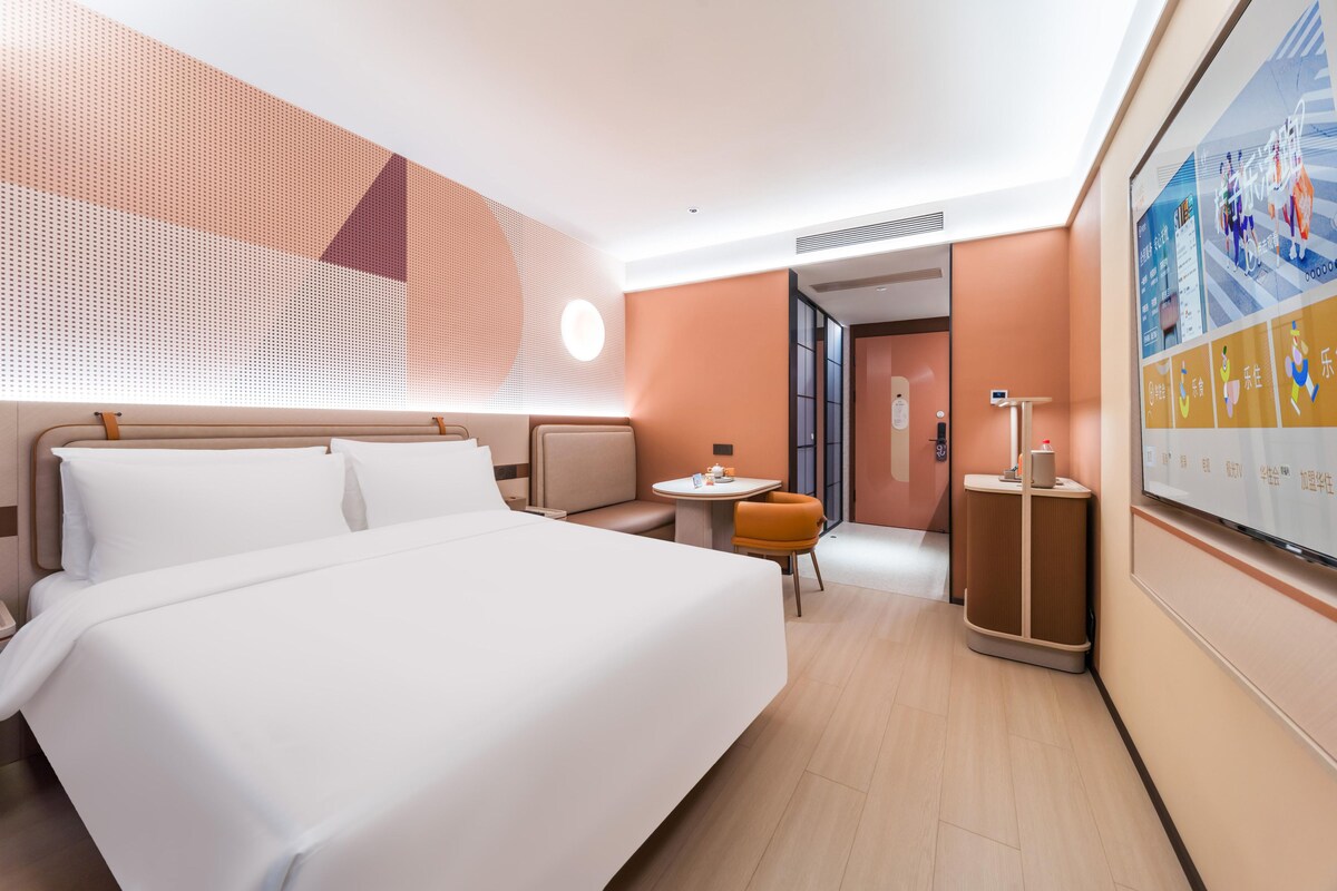 A hotel room with a large bed, a small table, a chair, and a TV screen displaying a sports event.
