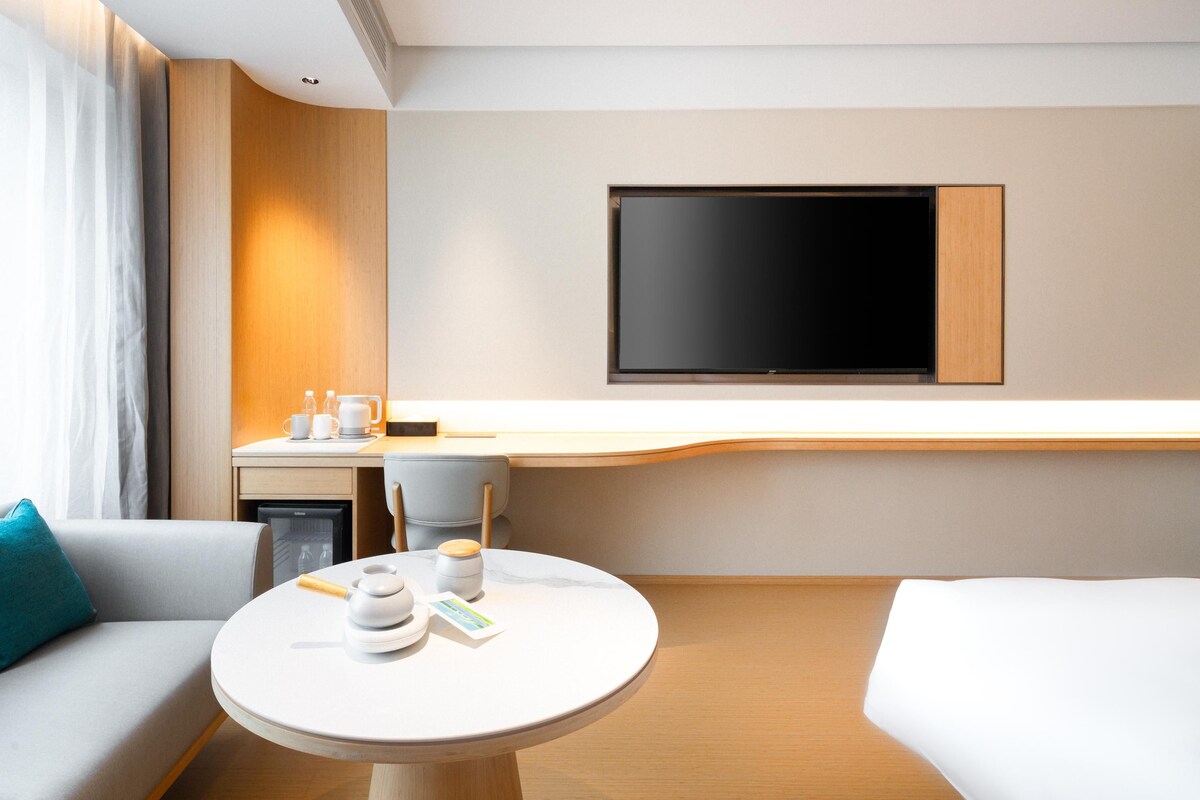 A modern hotel room with a sofa, a small round table with cups and a book, a desk with a chair, and a flat-screen TV mounted on the wall.