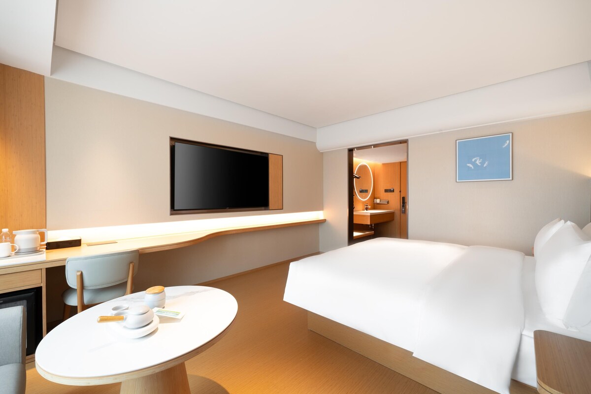 A modern hotel room with a large bed, a desk with a flat-screen TV, a round dining table, and a bathroom visible through an open door.