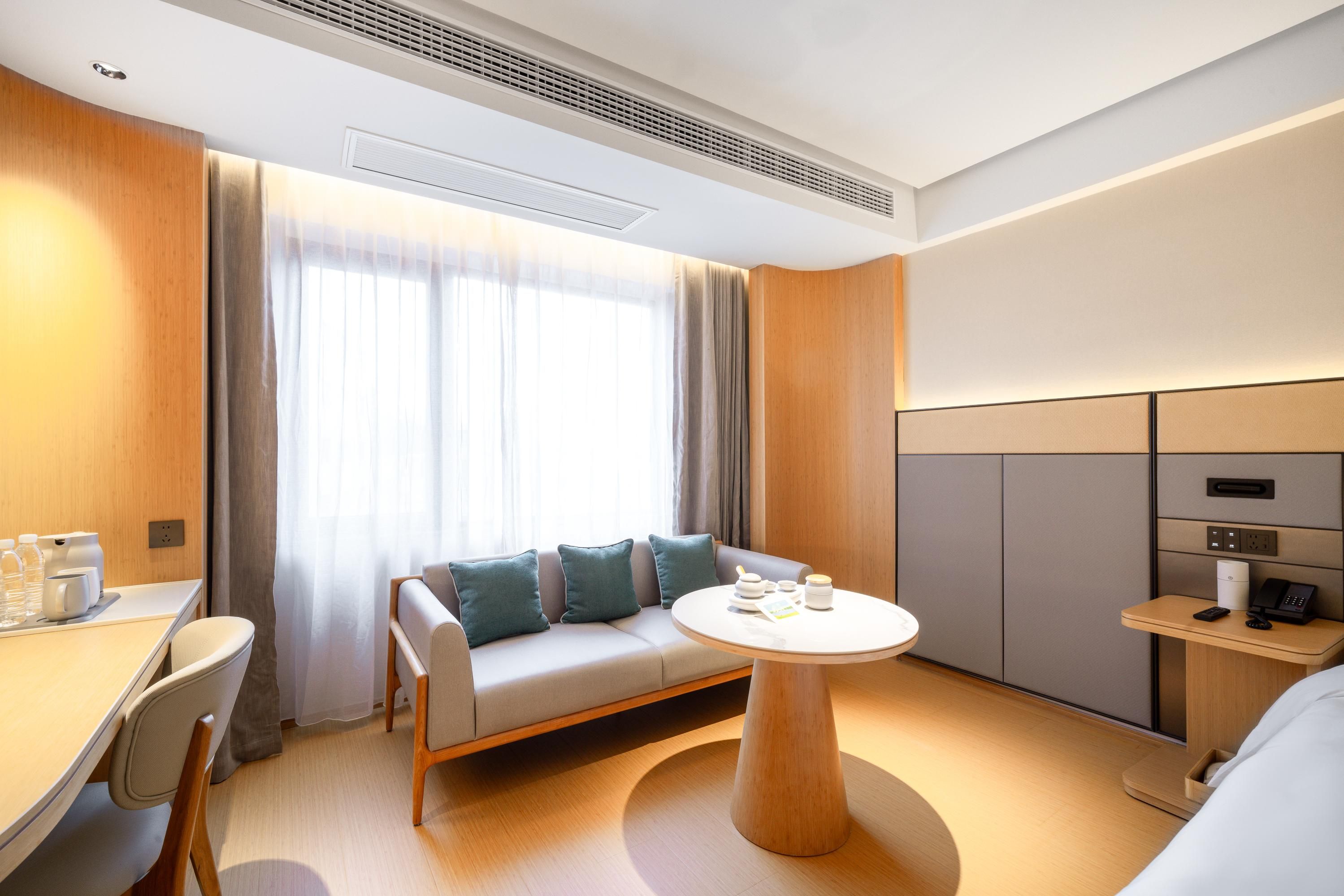 A modern hotel room with a sofa, a round table, a desk with chairs, and a bedside table with a phone and remote control.