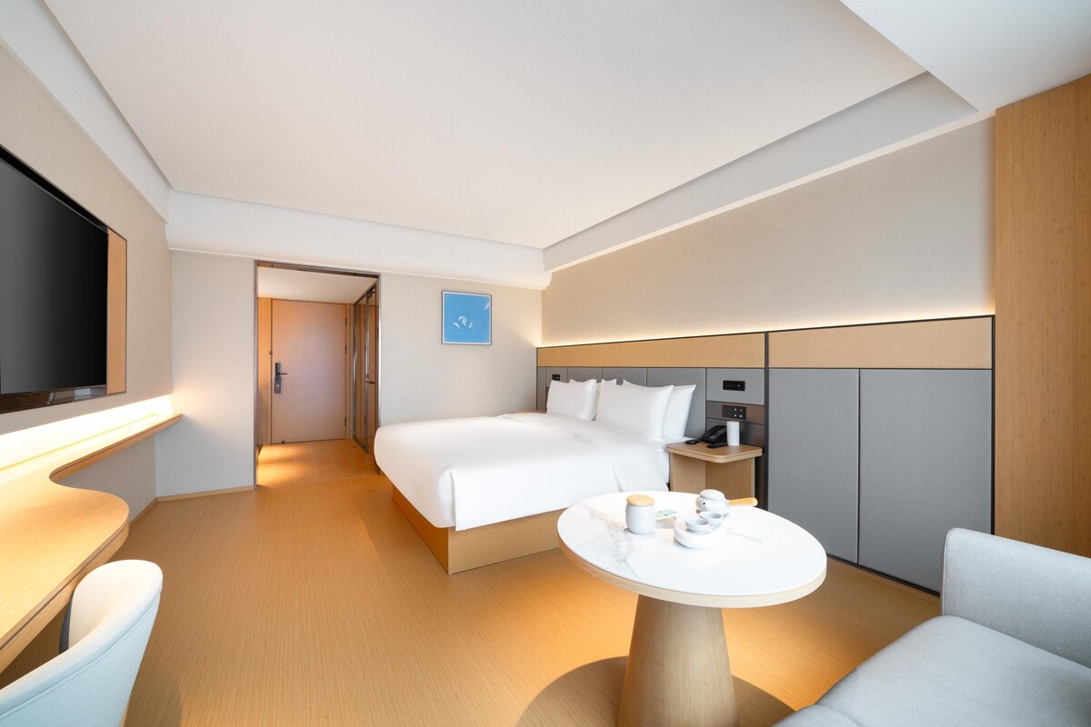 A modern hotel room with a bed, bedside table, and a small round table with a lamp.
