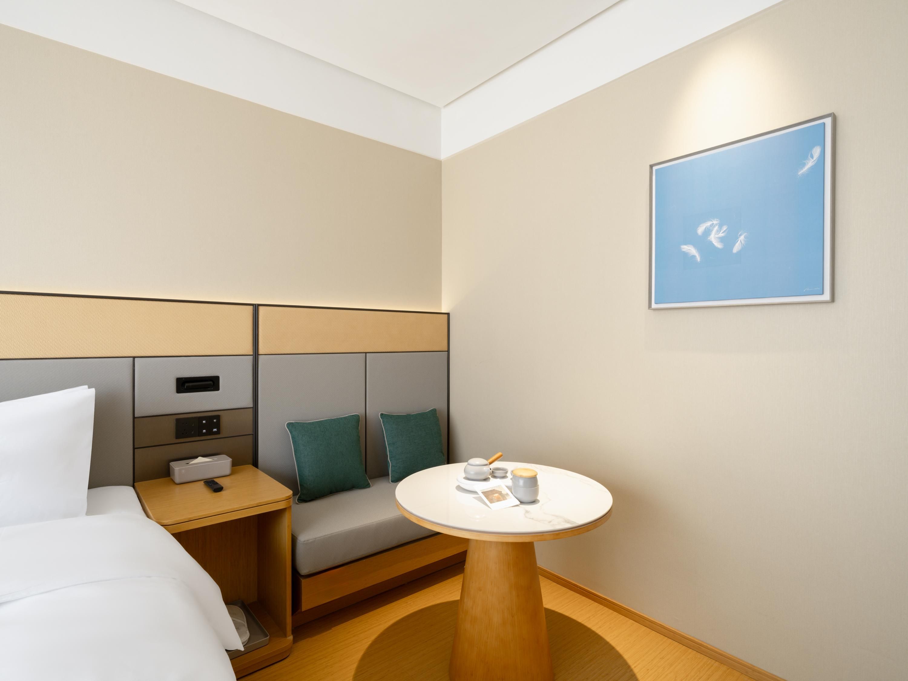 A hotel room with a bed, a bedside table with a lamp, a small round table with a cup and a remote control, a sofa with cushions, and a framed picture on the wall.
