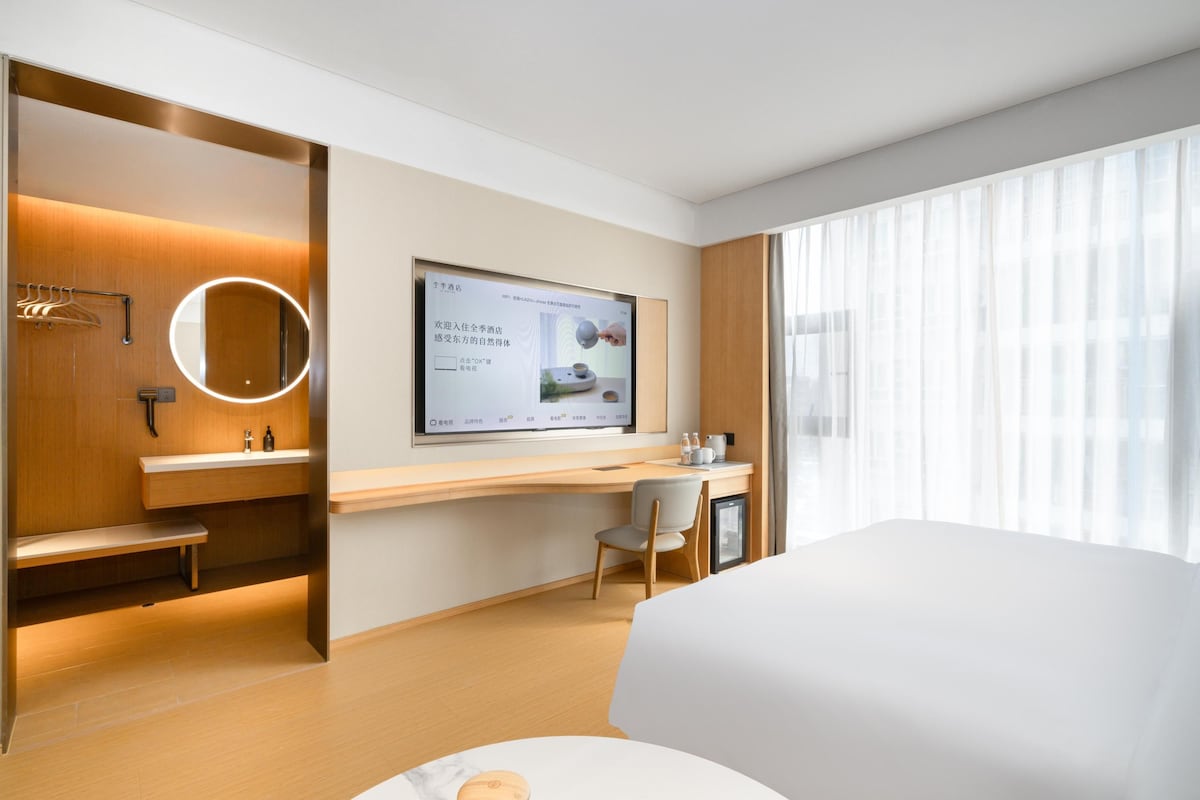 A modern hotel room with a large bed, a desk with a chair, a round mirror, and a TV mounted on the wall.