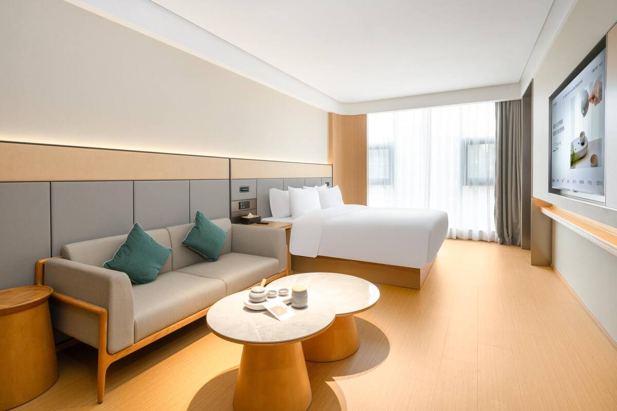 A modern hotel room with a sofa, a bed, a round coffee table, and a TV mounted on the wall.