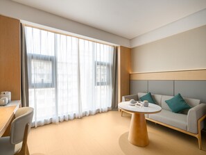 Una habitación de hotel moderna con un ventanal grande, una mesa redonda, un sofá con cojines y una mesita de madera con una tetera.