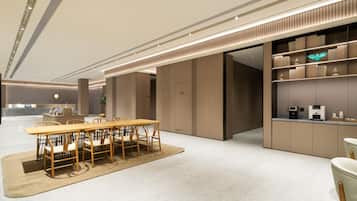 A modern hotel lobby with a reception desk, a dining area with a wooden table and chairs, and a kitchen area with shelves and appliances.