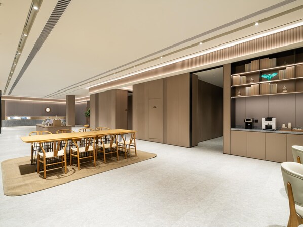 Un moderno vestíbulo de hotel con una recepción, un comedor con una mesa y sillas de madera, y una cocina con estantes y electrodomésticos.