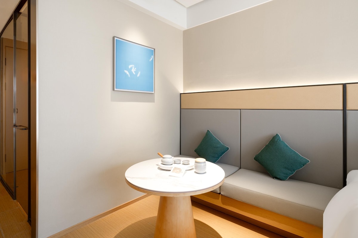 Una habitación de hotel moderna con una mesa redonda, un sofá con cojines azul turquesa y una obra de arte enmarcada en la pared.
