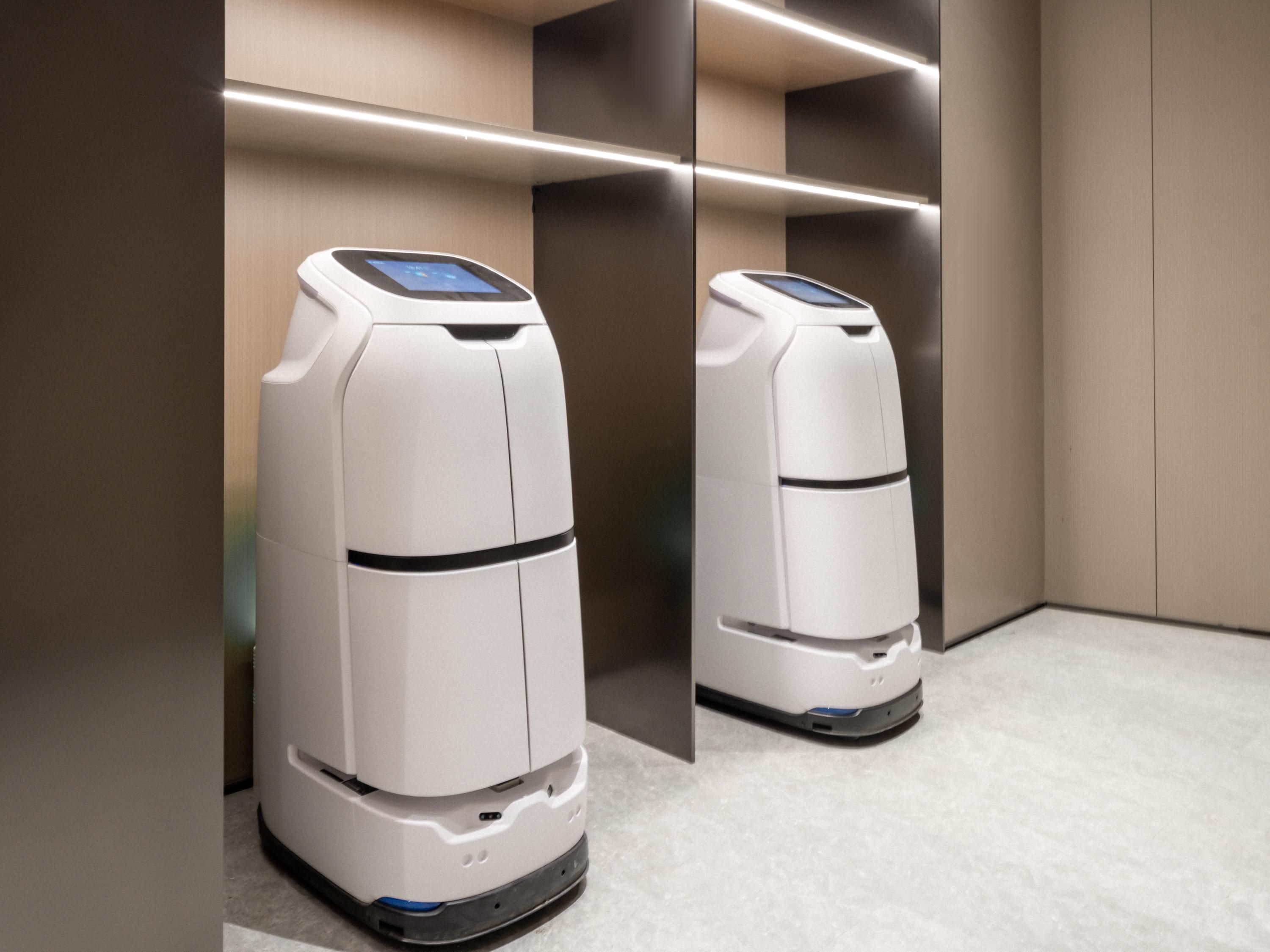 Two white robotic vacuum cleaners in a modern, empty elevator with a light brown wall and a grey floor.