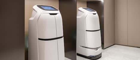 Dos aspiradoras robot blancas en un ascensor moderno y vacío, con una pared marrón claro y un piso gris.