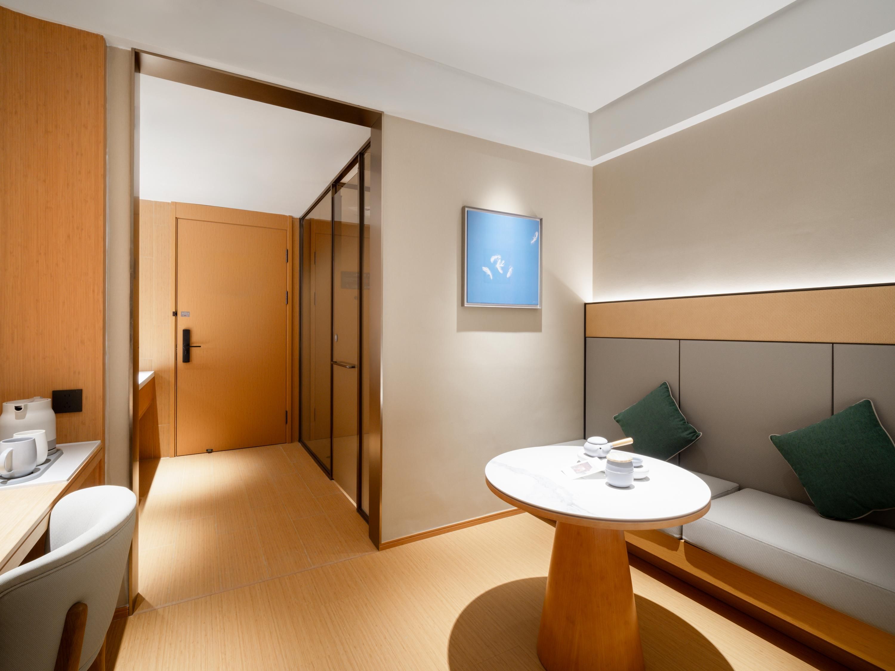 Une chambre d’hôtel moderne avec un plancher en bois, une table ronde au plateau blanc, un canapé avec des coussins verts, une porte en bois et un tableau encadré au mur.