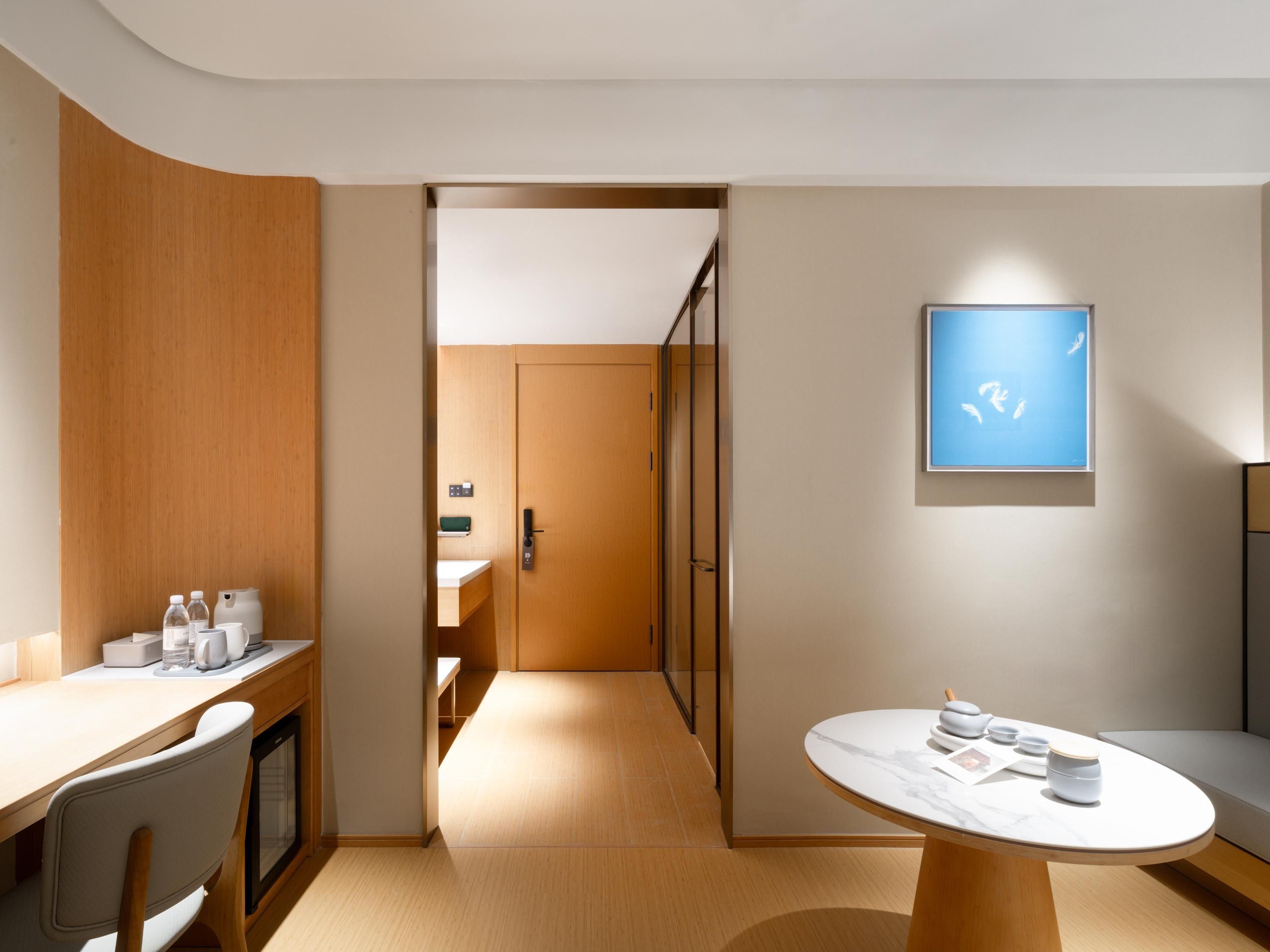A modern hotel room with a wooden desk, a round table with tea sets, a sofa, and a framed picture on the wall.