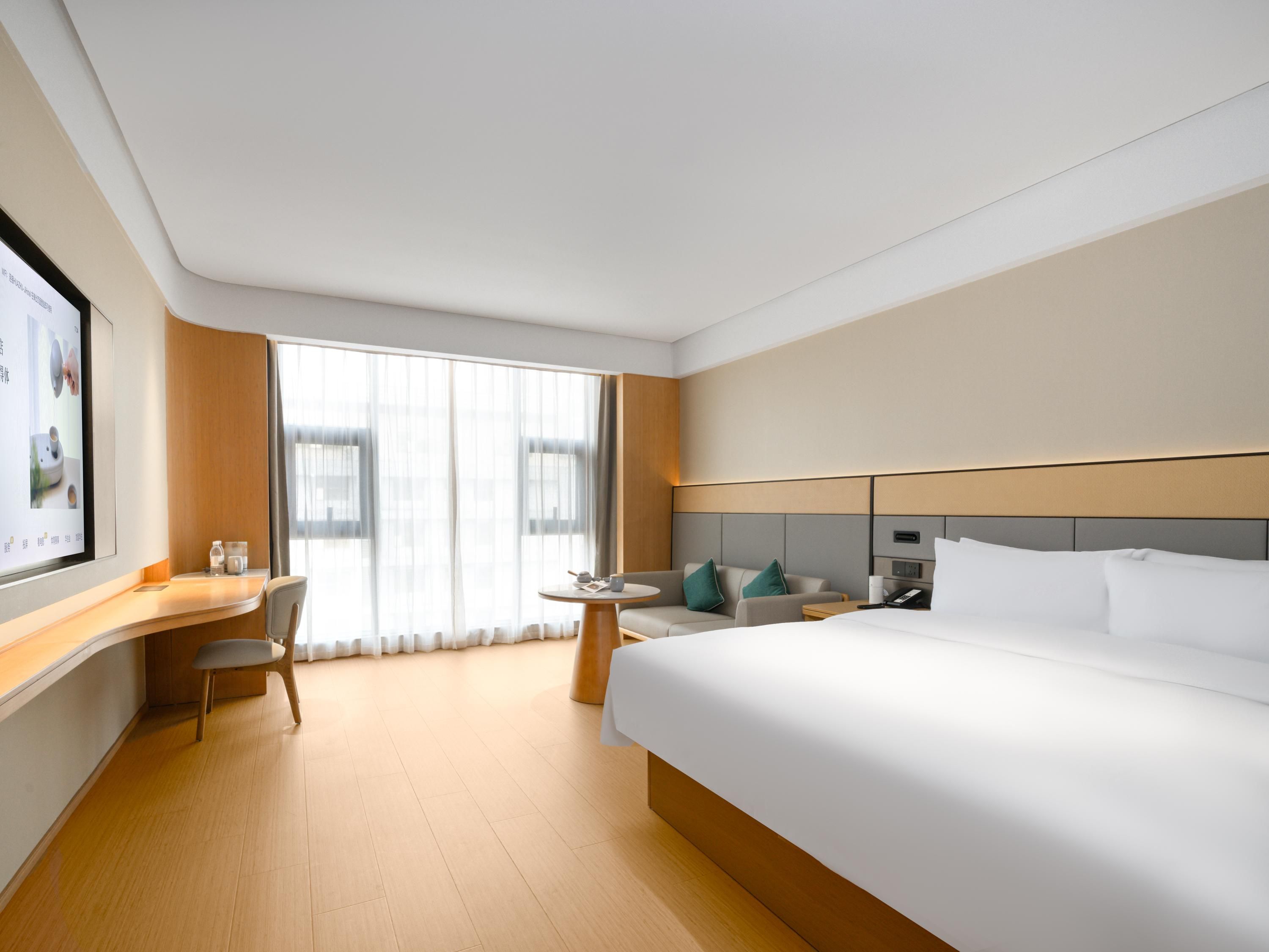 A hotel room with a large bed, a desk with a chair, a small table, and a large window with curtains.