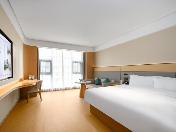 Una habitación de hotel con una cama grande, un escritorio con silla, una mesita pequeña y un ventanal con cortinas.