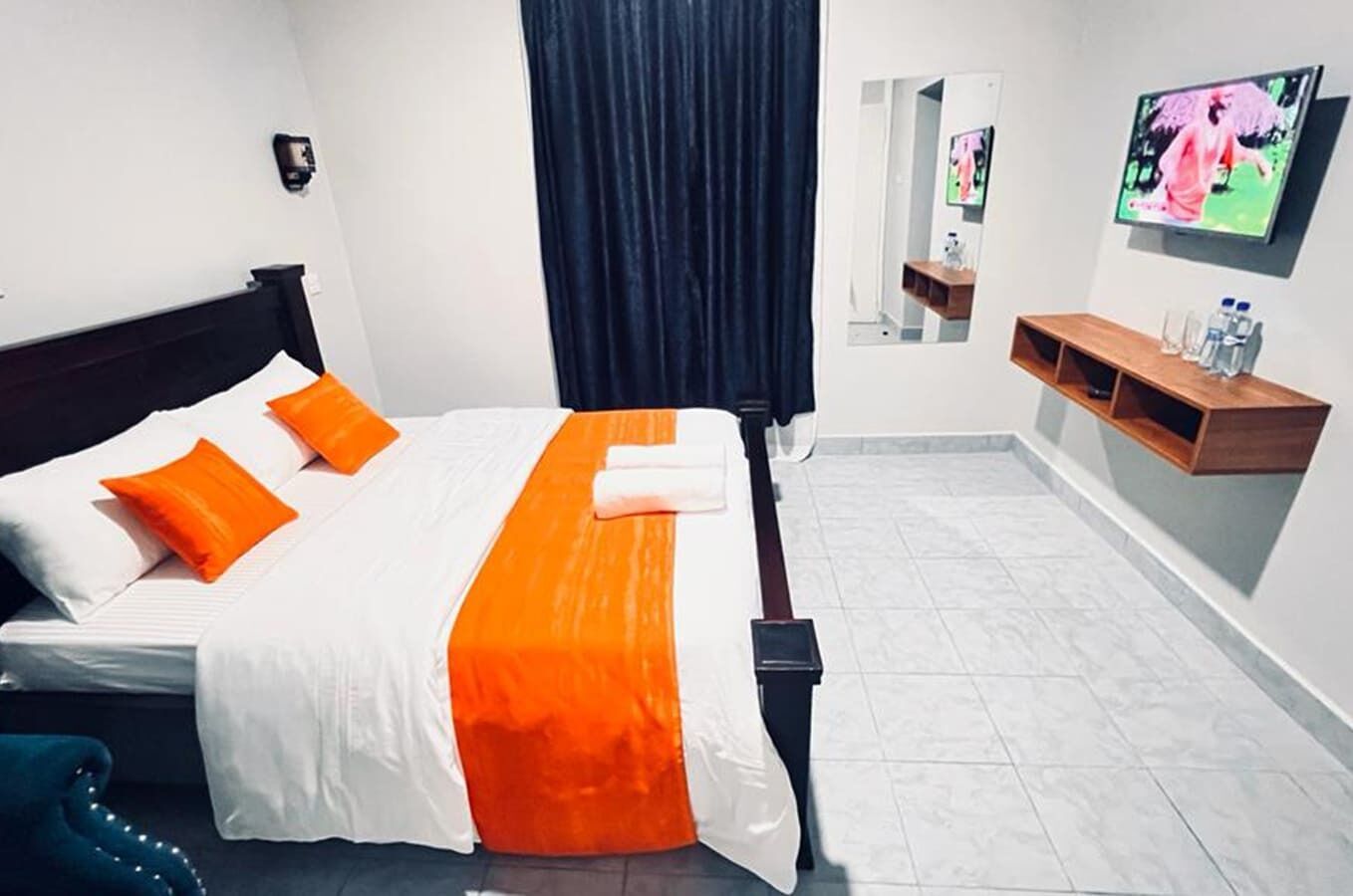 A hotel room with a bed, orange bedding, a TV on the wall, and a small shelf with toiletries.