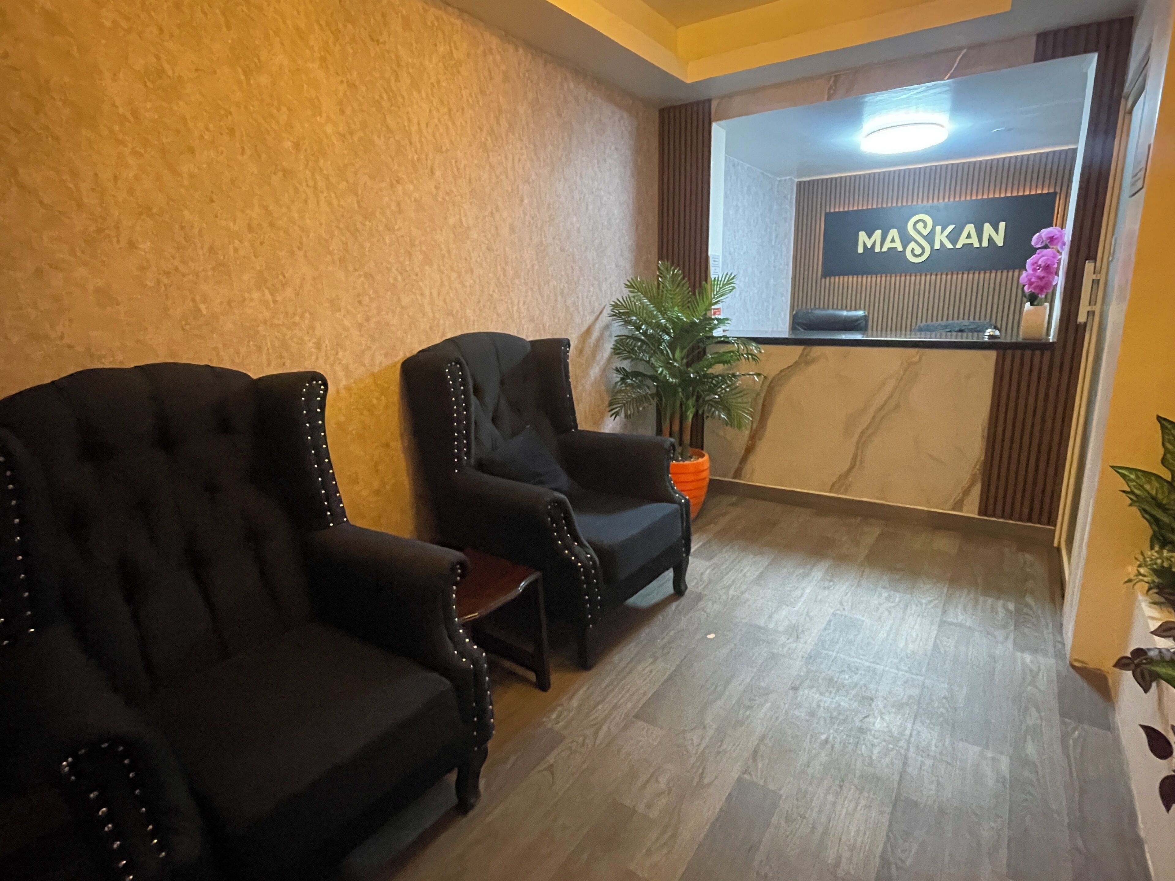 Un área de recepción con dos sillones tapizados en negro, un mostrador de recepción con el letrero 'MASKAN' y una planta en maceta.
