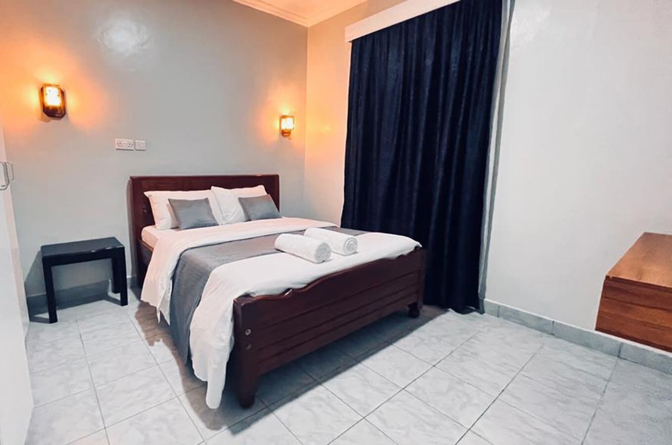 A neatly made bed with a wooden headboard, a small bedside table, and a dark curtain covering a window.