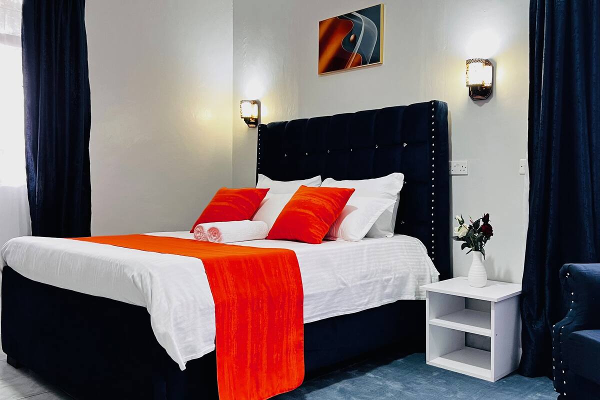 Une chambre d’hôtel avec un grand lit, du linge de lit blanc, des oreillers rouges et blancs, une table de chevet blanche ornée d’un vase de fleurs et des rideaux foncés.