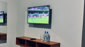 Un téléviseur fixé au mur, diffusant un programme sportif, une étagère en bois en dessous avec une télécommande et des bouteilles d’eau, et un miroir qui reflète l’image du téléviseur.