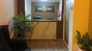Un espace d’accueil comprenant un comptoir de réception, une plante en pot et un panneau indiquant « MASKAN ».