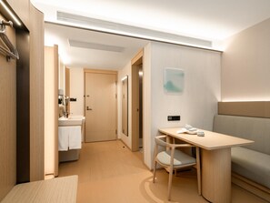 A modern hotel room with a wooden floor, a small desk with a chair, a sofa, a mirror, and a bathroom with a sink and towel. - Ji Hotel Guangzhou Baiyun Airport Renhe Metro (Guangzhou)