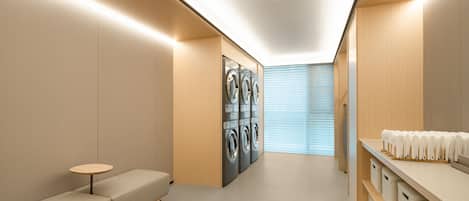 A modern laundry room with built-in washers and dryers, a seating area, and storage shelves.