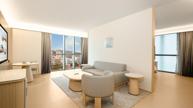 A modern living room with a sofa, coffee table, and side tables, large windows with curtains, and a view of a cityscape. - Ji Hotel Guangzhou Baiyun Airport Renhe Metro (Guangzhou)