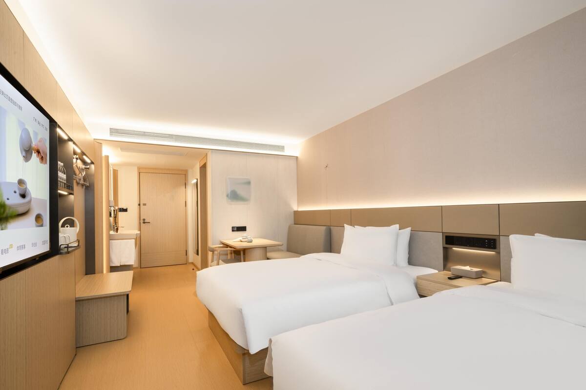 Habitación de hotel con dos camas, una mesita, una mesilla de noche, un televisor y un baño visible a través de una puerta abierta.