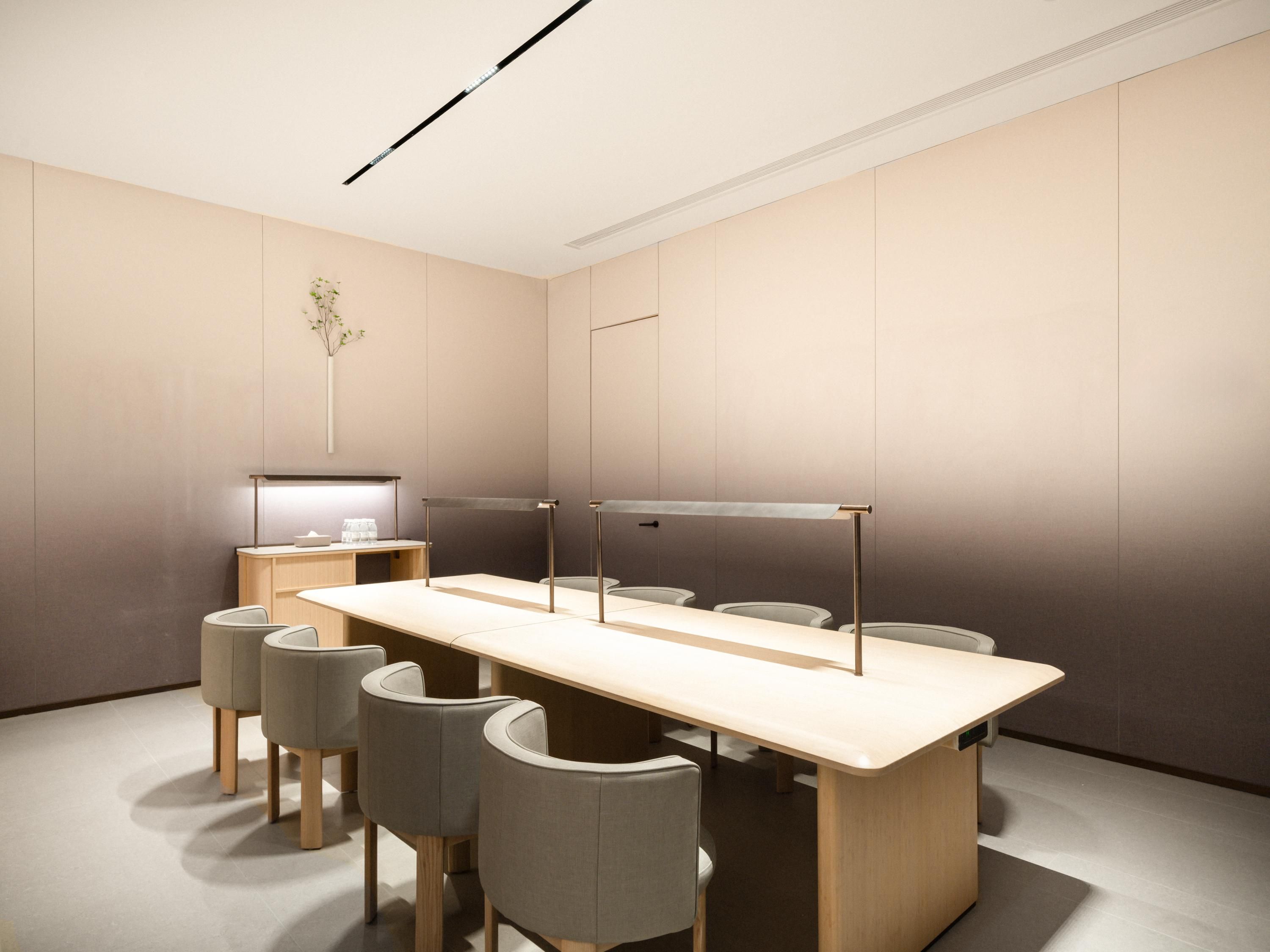 Uma sala de conferências moderna com uma mesa comprida, seis cadeiras, uma pequena escrivaninha com uma luminária e uma planta fixada na parede.